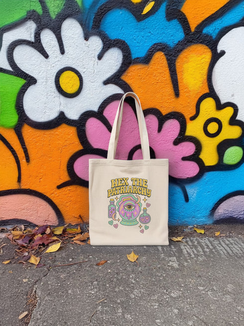 Canvas cotton tote bag with bold “Hex the Patriarchy” design printed on the front, featuring a dripping purple eye in a crystal ball surrounded by pastel potion bottles and hearts. Bag is available in black or natural cotton with long canvas handles.