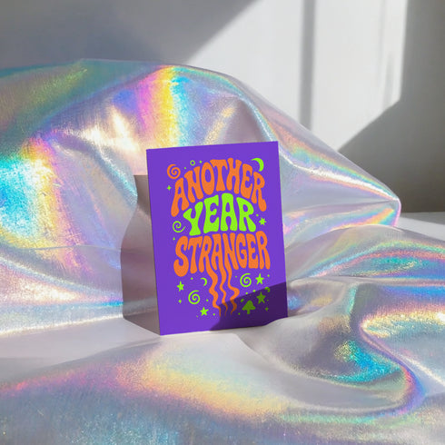 A purple greeting card with the text "Another Year Stranger" is displayed on a shiny, iridescent fabric surface.