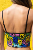 A sleeveless women's camisole top featuring a pointed handkerchief hemline often called a bandana top style. The fabric displays a vibrant all-over psychedelic print of distorting and melting smiley faces mixed with melting skulls. The color palette includes bright neon yellow, electric blue, hot pink, and lime green against a multi-colored background. The top features thin black adjustable spaghetti straps and black trim along the straight neckline. The fit is cropped and form-fitting.