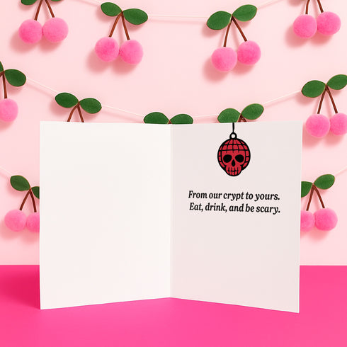 A 5x7 inch greeting card with a vibrant pink background and red retro text reading 'HOLIDAY IN THE CRYPT'. The illustration shows a male and female punk rock skeleton dancing under a red skull disco ball. The female skeleton has a mohawk and wears a black dress. The male skeleton has a mohawk and wears a black jacket with a red vest. Inside, the message reads, 'From our crypt to yours. Eat, drink, and be scary.' The style is a graphic, punk aesthetic.