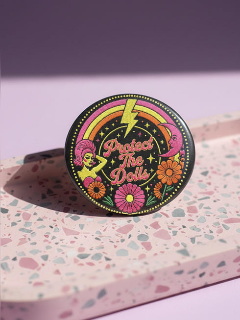 A circular black pinback button featuring the slogan Protect The Dolls in retro script. The design utilizes a high-fidelity trompe-l'œil print style that mimics the texture of raised thread embroidery with satin-stitch detailing. The artwork features vibrant hot pink, rust orange, and golden yellow motifs including drag queens with bouffant hair, 70s style daisies, lightning bolts, and a crescent moon with a face. The surface is smooth and glossy, creating a faux-textured illusion.