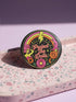 A circular black pinback button featuring the slogan Protect The Dolls in retro script. The design utilizes a high-fidelity trompe-l'œil print style that mimics the texture of raised thread embroidery with satin-stitch detailing. The artwork features vibrant hot pink, rust orange, and golden yellow motifs including drag queens with bouffant hair, 70s style daisies, lightning bolts, and a crescent moon with a face. The surface is smooth and glossy, creating a faux-textured illusion.