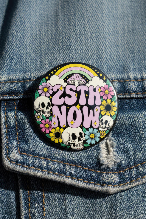 A round pinback button featuring a solid black background covered in a retro pastel colored design. The center reads 25TH NOW in a groovy bubbly font using pale pink and mint green referencing the 25th amendment. Surrounding the text is a maximalist cluster of cute but edgy motifs including smiling white skulls, pastel rainbows, pale yellow crescent moons, daisies, and a pink and white spotted mushroom. The button has a glossy laminate finish and a secure metal safety pin backing.