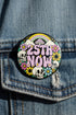 A round pinback button featuring a solid black background covered in a retro pastel colored design. The center reads 25TH NOW in a groovy bubbly font using pale pink and mint green referencing the 25th amendment. Surrounding the text is a maximalist cluster of cute but edgy motifs including smiling white skulls, pastel rainbows, pale yellow crescent moons, daisies, and a pink and white spotted mushroom. The button has a glossy laminate finish and a secure metal safety pin backing.