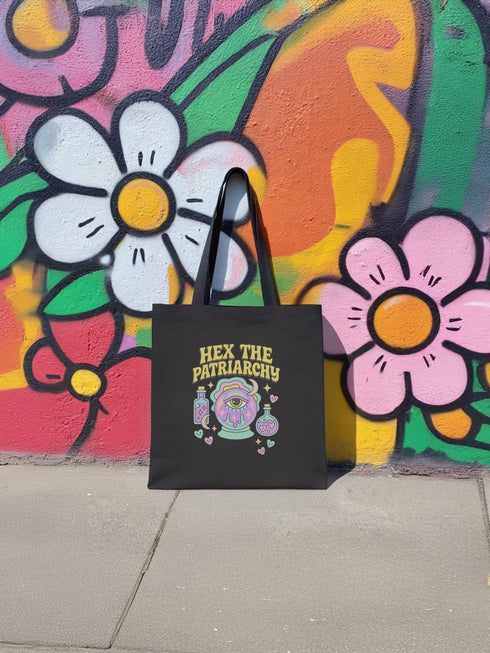 Canvas cotton tote bag with bold “Hex the Patriarchy” design printed on the front, featuring a dripping purple eye in a crystal ball surrounded by pastel potion bottles and hearts. Bag is available in black or natural cotton with long canvas handles.