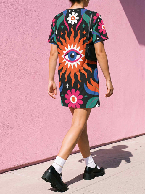 A short-sleeve, above-the-knee t-shirt dress with a straight, structured fit. The dress has a true black background featuring a bold, vibrant graphic pattern. The center displays a large blue and white evil eye surrounded by a bright orange wavy sunburst with a white jagged border. Above and below the eye are bright pink daisy flowers with orange centers, scattered white four-petal stars, and wavy blue and teal green leaf shapes. The fabric is thick, smooth, and holds its clean shape.