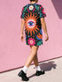 A short-sleeve, above-the-knee t-shirt dress with a straight, structured fit. The dress has a true black background featuring a bold, vibrant graphic pattern. The center displays a large blue and white evil eye surrounded by a bright orange wavy sunburst with a white jagged border. Above and below the eye are bright pink daisy flowers with orange centers, scattered white four-petal stars, and wavy blue and teal green leaf shapes. The fabric is thick, smooth, and holds its clean shape.