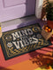 A rectangular floor mat with a dark charcoal black background. The center features the phrase 'MIND THE VIBES' in stylized, wavy cream and golden-mustard retro typography. The text is surrounded by symmetrical motifs: a golden evil eye with sun rays on the top left, a golden crescent moon with a four-point star on the top right, and two cream daisy flowers with golden stems at the bottom corners. The bottom edge has a wavy golden checkerboard pattern. A faux-stitch border frames the art.