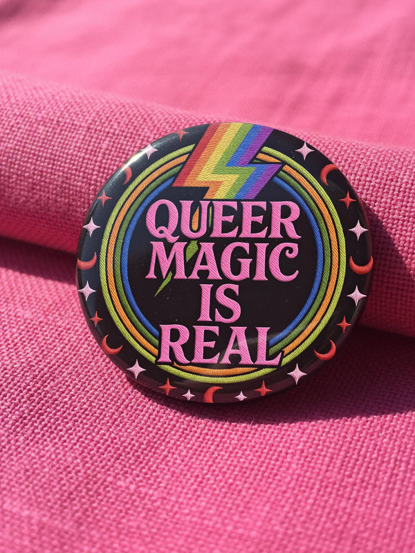 A round metal pinback button badge featuring a black background with the words QUEER MAGIC IS REAL in capital pink retro serif typography. A bright rainbow colored lightning bolt strikes from the top center, surrounded by concentric rainbow rings in red, orange, yellow, green, blue, and purple. The design is accented with small pink four-point stars and red crescent moons. The artwork features a realistic 2-D faux embroidery texture. The back has a silver metal safety pin clasp fastening.