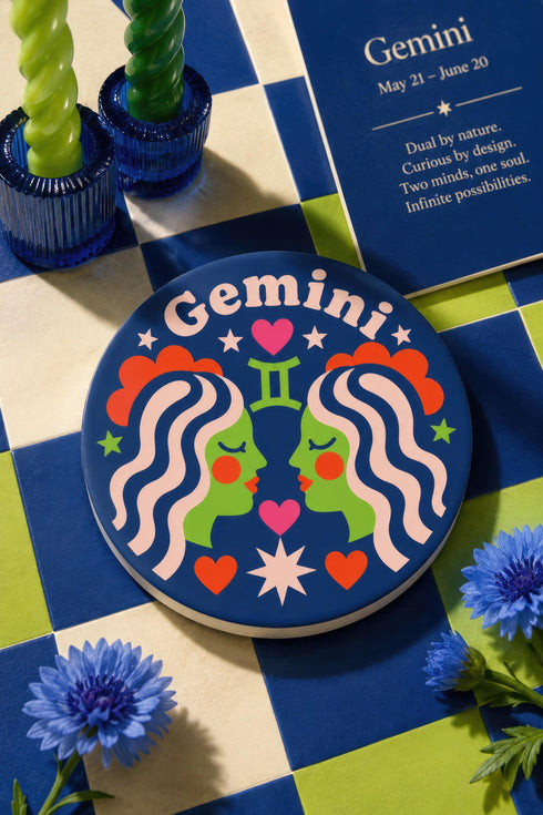 A close-up photograph of a round, glossy ceramic coaster featuring a bold retro pop art design. The background is a deep royal blue. In the center are two symmetrical profile faces of twins looking at each other, featuring bright green skin, closed eyes, and red cheeks. They have wavy white and pale pink hair with red accents. The word Gemini is arched at the top in a retro white font. The design is decorated with a green Gemini symbol, pink and red hearts, and white starbursts.