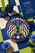 A close-up photograph of a round, glossy ceramic coaster featuring a bold retro pop art design. The background is a deep royal blue. In the center are two symmetrical profile faces of twins looking at each other, featuring bright green skin, closed eyes, and red cheeks. They have wavy white and pale pink hair with red accents. The word Gemini is arched at the top in a retro white font. The design is decorated with a green Gemini symbol, pink and red hearts, and white starbursts.