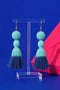 A pair of turquoise tassel earrings with blue tassels hanging from a clear stand against a blue background.