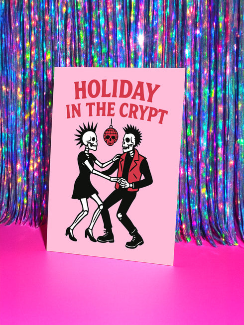A 5x7 inch greeting card with a vibrant pink background and red retro text reading 'HOLIDAY IN THE CRYPT'. The illustration shows a male and female punk rock skeleton dancing under a red skull disco ball. The female skeleton has a mohawk and wears a black dress. The male skeleton has a mohawk and wears a black jacket with a red vest. Inside, the message reads, 'From our crypt to yours. Eat, drink, and be scary.' The style is a graphic, punk aesthetic.