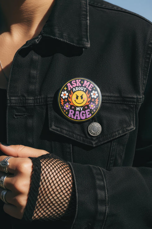 A round glossy metal pinback button featuring a flat 2D print design on a solid black background. In the center is an angry yellow smiley face surrounded by orange and red flames. Above the smiley face, pink text reads ASK ME and white text reads ABOUT. Below the face, white text reads MY and large purple text reads RAGE. The design is accented with two white daisies, two purple flowers, pink and orange four-point stars, and orange lightning bolts framing the outer edges.