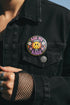 A round glossy metal pinback button featuring a flat 2D print design on a solid black background. In the center is an angry yellow smiley face surrounded by orange and red flames. Above the smiley face, pink text reads ASK ME and white text reads ABOUT. Below the face, white text reads MY and large purple text reads RAGE. The design is accented with two white daisies, two purple flowers, pink and orange four-point stars, and orange lightning bolts framing the outer edges.
