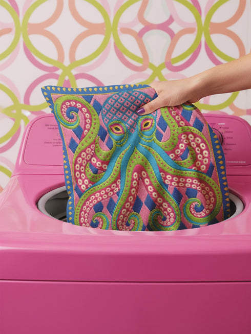 A square decorative throw pillow cover featuring a highly symmetrical bright octopus design. The octopus has a turquoise blue body with a diamond patterned head and eight curled tentacles detailed in bright pink and lime green. The background is a repeating diamond argyle pattern in royal blue and light pink. The artwork is a flat two dimensional print that mimics the textured look of intricate crewelwork embroidery. The pillow features a hidden zipper on the bottom edge. No insert included.