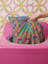 A square decorative throw pillow cover featuring a highly symmetrical bright octopus design. The octopus has a turquoise blue body with a diamond patterned head and eight curled tentacles detailed in bright pink and lime green. The background is a repeating diamond argyle pattern in royal blue and light pink. The artwork is a flat two dimensional print that mimics the textured look of intricate crewelwork embroidery. The pillow features a hidden zipper on the bottom edge. No insert included.