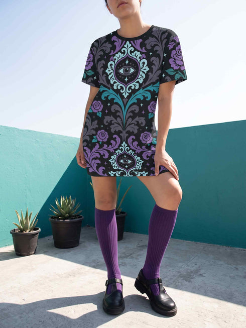 Cotton t-shirt dressA short-sleeve structured t-shirt dress with a black background. The dress features a bold, vertically stacked repeating graphic print. The design includes intricate damask patterns in teal and grey, purple roses with teal leaves, and a central evil eye motif set inside an ornate frame with small four-point stars. The heavyweight fabric holds a clean, straight cut above the knee with zero drape, maintaining a sculptural, slightly boxy silhouette.