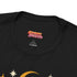 A unisex short sleeve graphic t shirt in dark charcoal black featuring a highly detailed symmetrical medallion print on the chest. In the center, the word RESIST is printed in a bold yellow gothic blackletter font. Two coiled white snakes with fine black scale details frame the text on the left and right. Above the text, a yellow crescent moon is flanked by white and yellow starbursts. Beneath, three yellow marigold flowers sit atop white stems and leafy foliage.