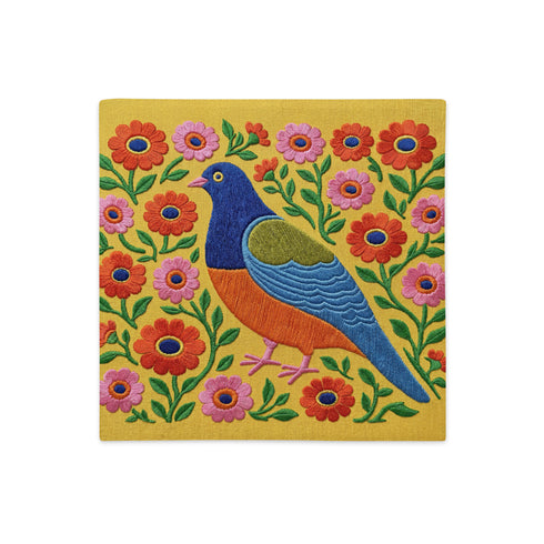 Square faux embroidery throw pillow cover with a large stylized folk art bird facing left. The bird has a cobalt blue head, orange belly, light blue tail, and pink legs, set on a bright mustard yellow background scattered with pink and red daisy flowers on trailing green vines. The smooth, linen-feel polyester uses a printed trompe loeil effect to mimic raised thread needlework. Bold maximalist folk art bird pillowcase and cottagecore cushion cover for eclectic, colorful, dopamine decor homes.