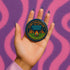 Round glossy pinback button measuring 2.25 inches featuring a playful camp-themed design with the phrase “Camp Counselor for the Gender Crisis.” The artwork includes two orange tents, a rainbow campfire, stars, and green grass. Bottom text reads “Cabin Queer, Unit Psychedelic.” Made from durable tinplate with a safety pin back and coated in a UV and scratch-resistant mylar finish. Bold and colorful, perfect for jackets, bags, or pride outfits.