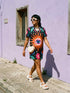 A short-sleeve, above-the-knee t-shirt dress with a straight, structured fit. The dress has a true black background featuring a bold, vibrant graphic pattern. The center displays a large blue and white evil eye surrounded by a bright orange wavy sunburst with a white jagged border. Above and below the eye are bright pink daisy flowers with orange centers, scattered white four-petal stars, and wavy blue and teal green leaf shapes. The fabric is thick, smooth, and holds its clean shape.