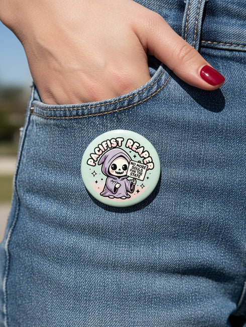 Round pinback button featuring a pastel mint green background with a cartoon grim reaper character in a lavender hood holding a protest sign. The design includes bold black outlined lettering reading “Pacifist Reaper” above the figure and small sparkle accents surrounding it. The finish is glossy and smooth, with a printed illustration style on a metal pin badge surface.