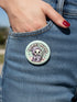 Round pinback button featuring a pastel mint green background with a cartoon grim reaper character in a lavender hood holding a protest sign. The design includes bold black outlined lettering reading “Pacifist Reaper” above the figure and small sparkle accents surrounding it. The finish is glossy and smooth, with a printed illustration style on a metal pin badge surface.