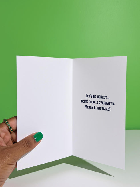 A vertical 5x7 holiday card featuring a retro tattoo style illustration of Krampus in pastel pink, teal, and black ink on shimmery silver pearlescent paper. The central horned demon has a long tongue and is surrounded by vintage rotary phones, daggers, candy canes, and skull gifts. Top text asks Have You Been Good while the bottom banner reads Krampus Is Calling. The interior text reads Let's be honest... Being good is overrated. Merry Christmas.