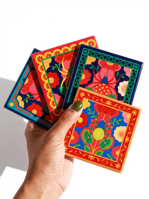 A set of four square coasters featuring a glossy glazed finish and vibrant insect illustrations. Each coaster measures approximately four by four inches. The designs showcase symmetrical beetles and spiders in bold red, blue, and orange colors, framed by intricate floral and geometric borders in a maximalist folk art style. Available in ceramic with cork backing or hardboard MDF. The edges are smooth. The designs are vivid and highly saturated, designed to provide grip and protect furniture.