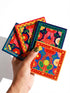 A set of four square coasters featuring a glossy glazed finish and vibrant insect illustrations. Each coaster measures approximately four by four inches. The designs showcase symmetrical beetles and spiders in bold red, blue, and orange colors, framed by intricate floral and geometric borders in a maximalist folk art style. Available in ceramic with cork backing or hardboard MDF. The edges are smooth. The designs are vivid and highly saturated, designed to provide grip and protect furniture.