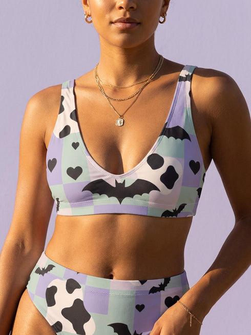 A two piece recycled high waisted swimsuit with a pastel goth checkered print in light lavender purple, pale mint green, and white squares with black cow print spots. The design features small black hearts and silhouetted bats with outspread wings printed throughout. The athletic style crop top has a deep scoop neck and matching thick straps, while the double layered bottoms have a supportive high waistband and cheeky cut with a solid black inner lining.
