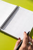 A person's hand holding a black pen is seen writing on a white calendar page with a black spiral binding, set against a bright green background.