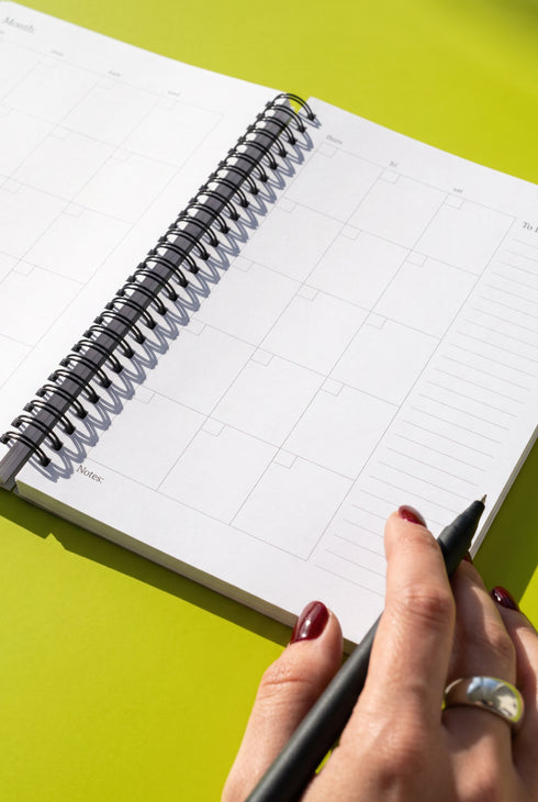 A person's hand holding a black pen is seen writing on a white calendar page with a black spiral binding, set against a bright green background.