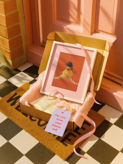 A fine art print of a rubber duck dressed like Alex from A Clockwork Orange, featuring a black bowler hat, blunt bob, and white suspenders. The duck floats in orange-tinged bubbles and is framed in white on a glossy pink tile bathroom shelf.