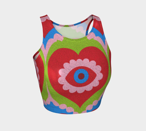 A sleeveless scoop neck crop top featuring a bold all-over printed design. The central graphic is a large bright red heart enclosing a scalloped pink flower shape and a blue evil eye motif in the middle. The background surrounds the heart with mirrored concentric wavy arches and scalloped borders in vibrant shades of lime green, pink, blue, and red. The fitted garment has a smooth athletic texture. Available in a standard matte performance knit or a sparkly silver glitter foil finish.