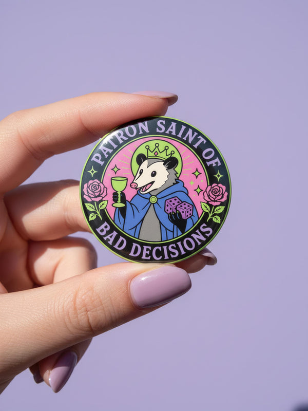 A round glossy metal pinback button featuring a flat 2D print illustration of an opossum wearing a blue cloak and a green crown. The opossum holds a neon green goblet in one paw and two purple dice in the other. A bright pink circle sits behind the opossum, surrounded by a black outer ring. Purple text reads PATRON SAINT OF BAD DECISIONS curved around the edge. Two pink roses with green leaves are on the sides. The design uses bold black outlines and bright neon colors.