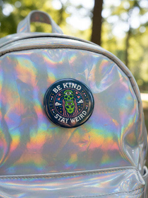 A round 1.75 inch pinback button featuring a retro psychedelic design on a dark navy blue background. The text reads BE KIND STAY WEIRD in vintage cream lettering around the border. The center illustration depicts a neon green skull stacked on top of a neon green alien head. Both the skull and alien have opening third eyes on their foreheads. Pink wavy lines frame the faces. The background is decorated with small colorful mushrooms, hearts, stars, and clouds. Glossy finish.