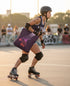 A woman rollerblading on a concrete surface, wearing a helmet and protective gear, with a purple bag featuring a graphic design attached to her arm.