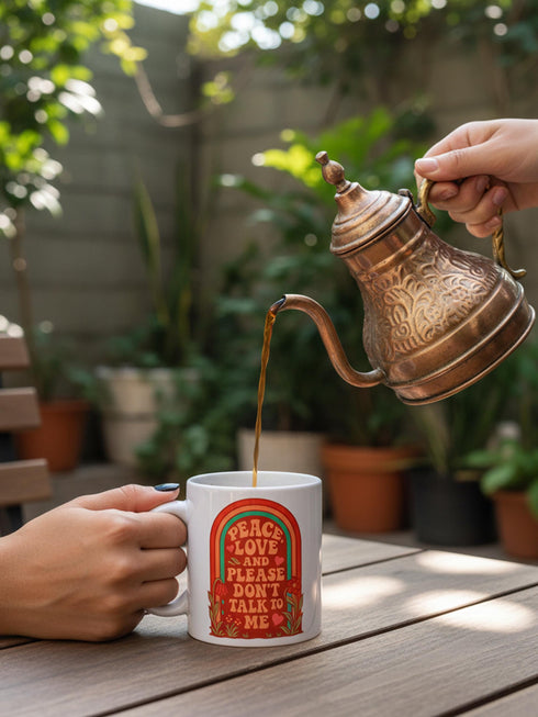 A glossy white porcelain ceramic mug featuring a vibrant maximalist design centered on the exterior. The artwork displays a retro 1970s-inspired rainbow arch composed of concentric bands in burnt orange, mustard yellow, and teal green. Inside the arch, stylized vintage bubble-serif typography in reddish-orange reads PEACE LOVE AND PLEASE DON'T TALK TO ME. The design evokes a groovy, hippie aesthetic mixed with sassy introvert humor.