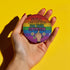 Round glossy pinback button measuring 2.25 inches with a bold rainbow pride flag background. Centered text reads “Still Not Asking For Your Opinion” in black, yellow, blue, and purple letters. Below the text is an illustrated hand with painted red nails giving the middle finger, surrounded by white daisies. The design is printed on tinplate with a scratch- and UV-resistant mylar coating. Button features a strong safety pin backing, perfect for bags, jackets, or pride outfits.