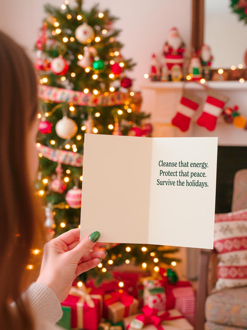 A detailed folk-art style illustration of a "Sage the Halls" holiday greeting card. The design features a bundle of sage smudge sticks tied with a red ribbon at the center of a dark green wreath. The wreath is accented with small crescent moons, stars, and smoke swirls. Below the wreath are two crossed matches with red tips. The card is printed on creamy beige paper with bold, dark green serif text. This funny spiritual and witchy Yule card is a unique pagan greeting for energy cleansing.