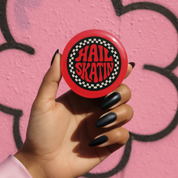 A round red pinback button with a glossy, scratch-resistant mylar face. The design features the phrase "HAIL SKATIN'" in a wavy, 70s psychedelic font in the center. This text is surrounded by a bold, black and white checkerboard pattern border, giving it a punk rock aesthetic. This piece of vintage-inspired flair is made of lightweight metal with a durable safety pin backing on the reverse. The button is a perfect for a denim jacket, backpack, or bag for fans of roller derby and skateboarding.