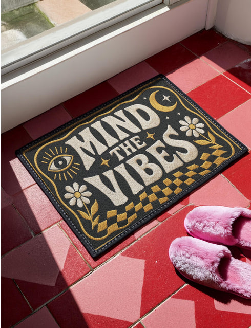 A rectangular floor mat with a dark charcoal black background. The center features the phrase 'MIND THE VIBES' in stylized, wavy cream and golden-mustard retro typography. The text is surrounded by symmetrical motifs: a golden evil eye with sun rays on the top left, a golden crescent moon with a four-point star on the top right, and two cream daisy flowers with golden stems at the bottom corners. The bottom edge has a wavy golden checkerboard pattern. A faux-stitch border frames the art.