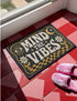 A rectangular floor mat with a dark charcoal black background. The center features the phrase 'MIND THE VIBES' in stylized, wavy cream and golden-mustard retro typography. The text is surrounded by symmetrical motifs: a golden evil eye with sun rays on the top left, a golden crescent moon with a four-point star on the top right, and two cream daisy flowers with golden stems at the bottom corners. The bottom edge has a wavy golden checkerboard pattern. A faux-stitch border frames the art.