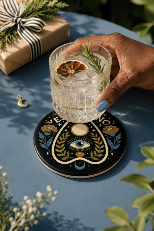 A close-up photograph of a glossy ceramic coaster featuring a Planchette occult design on a deep black background. The symmetrical graphic displays a central blue All-Seeing Eye with gold sunburst lines, topped by a white circle, and flanked by gold flowers with blue stems. At the top, the text "YES" is printed in gold on the left and "NO" on the right. Gold stars and crescent moons are scattered around the perimeter. The flat, round coaster has a highly visible high-gloss glaze.