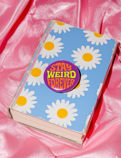 A round metal pin button with a glossy finish and a vibrant purple background. The phrase 'STAY WEIRD FOREVER' is written across the center in a wavy, psychedelic 70s-style font. The words 'STAY' and 'FOREVER' are a bold, bright orange, while the word 'WEIRD' is rendered in a contrasting, electric chartreuse lime green. The button has a smooth, scratch-resistant mylar surface covering the design. The pin is shown in its 2-inch and 3-inch variations, highlighting its circular shape and durable construction.