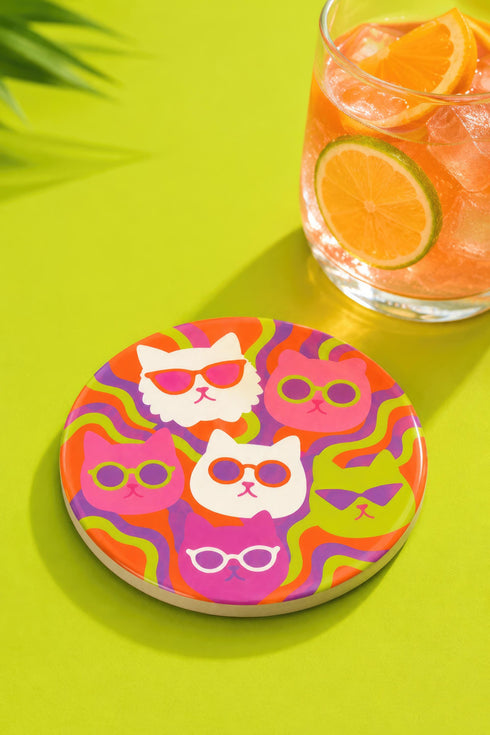 A close-up photograph of a glossy round ceramic coaster featuring a retro 70s psychedelic design. The artwork displays a cluster of six stylized cat face illustrations, each wearing a different pair of sunglasses including retro round and cat-eye shapes. The cats are colored in solid shades of white, hot pink, purple, and lime green. They are set against a vibrant background of wavy, swirling lines in bright orange, red, and purple. The top surface has a highly reflective clear glaze.