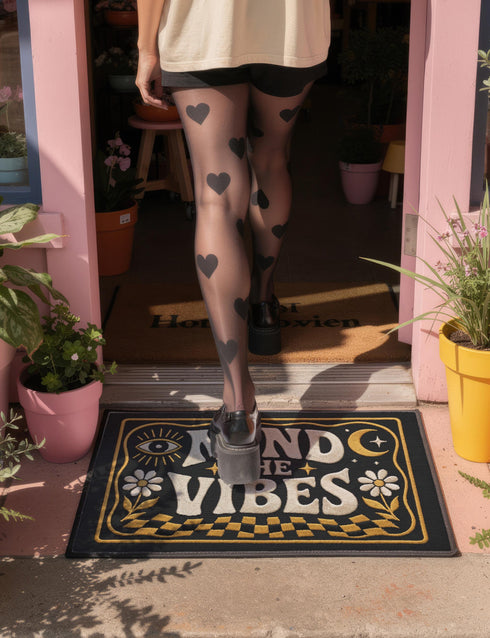 A rectangular floor mat with a dark charcoal black background. The center features the phrase 'MIND THE VIBES' in stylized, wavy cream and golden-mustard retro typography. The text is surrounded by symmetrical motifs: a golden evil eye with sun rays on the top left, a golden crescent moon with a four-point star on the top right, and two cream daisy flowers with golden stems at the bottom corners. The bottom edge has a wavy golden checkerboard pattern. A faux-stitch border frames the art.