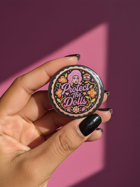 A circular black pinback button featuring the slogan Protect The Dolls in retro script. The design utilizes a high-fidelity trompe-l'œil print style that mimics the texture of raised thread embroidery with satin-stitch detailing. The artwork features vibrant hot pink, rust orange, and golden yellow motifs including drag queens with bouffant hair, 70s style daisies, lightning bolts, and a crescent moon with a face. The surface is smooth and glossy, creating a faux-textured illusion.